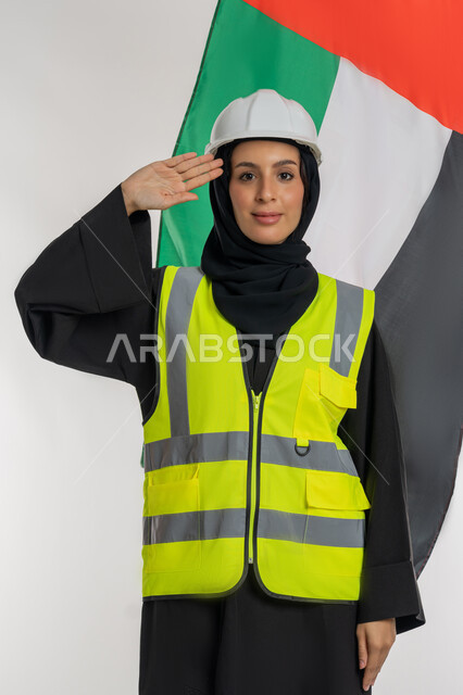 رفع علم الامارات عاليا، الاحتفال بيوم العلم الإماراتي 3 نوفمبر، الفخر والإعتزاز بالانتماء للوطن، ذكرى يوم الاستقلال 2 ديسمبر، بورتريه لمهندسة عربية خليجية اماراتية ترتدي سترة وخوذة الحماية تضع يدها على رأسها بإيماءات القاء التحية والاحترام، خلفية بيضاء