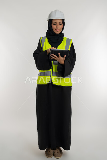 العمل في القطاع الهندسي، انجاز المهام عن بعد، إدارة المشاريع الهندسية, بورتريه  لمهندسة عربية خليجية اماراتية ترتدي سترة وخوذة الحماية تعمل على جهاز التابلت, شابة سعودية تقوم بدراسة أساسيات المشروع، صورة لكامل الجسم، خلفية بيضاء
