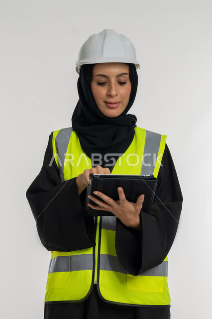 تعابير التركيز والانشغال، انجاز المهام عن بعد، إدارة المشاريع الهندسية، العمل في القطاع الهندسي, بورتريه  لمهندسة عربية خليجية اماراتية ترتدي سترة وخوذة الحماية تعمل على جهاز التابلت, شابة سعودية تقوم بدراسة أساسيات المشروع، خلفية بيضاء