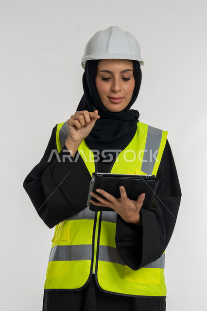 Gestures of focus and preoccupation, completing tasks remotely, managing engineering projects, working in the engineering sector, portrait of an Arab Gulf Emirati female engineer wearing a protective jacket and helmet working on a tablet, a young Saudi woman studying the basics of the project, white background