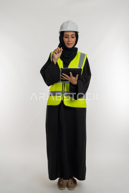 Completing tasks remotely, managing engineering projects, working in the engineering sector, portrait of an Arab Gulf Emirati female engineer wearing a protective jacket and helmet working on a tablet, a young Saudi woman studying the basics of the project, full body image, white background