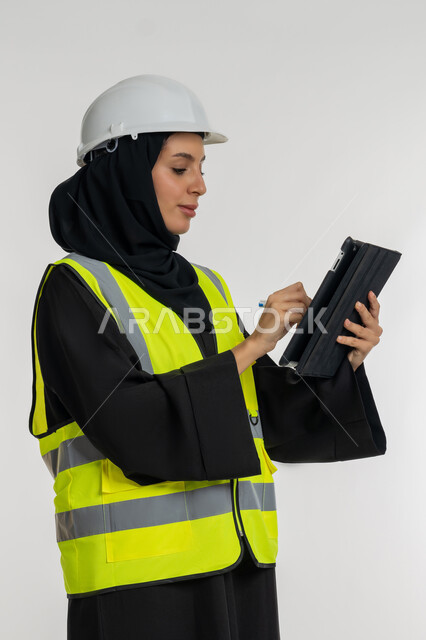 Engineering project management, working in engineering sector, completing tasks remotely, side portrait of an Arab Gulf Emirati female engineer wearing a protective jacket and helmet working on a tablet, a young Saudi woman studying the basics of the project, white background