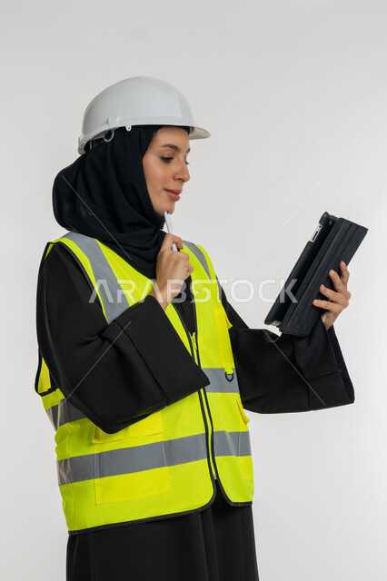ايماءات التركيز والانشغال، انجاز المهام عن بعد، إدارة المشاريع الهندسية، العمل في القطاع الهندسي, بورتريه من الجانب  لمهندسة عربية خليجية اماراتية ترتدي سترة وخوذة الحماية تعمل على جهاز التابلت, شابة سعودية تقوم بدراسة أساسيات المشروع، خلفية بيضاء
