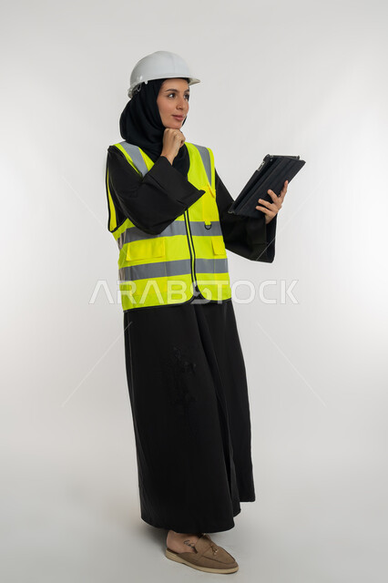 Studying project basics, working in engineering sector, engineering project management, portrait of an Arab Gulf Emirati female engineer wearing a protective jacket and helmet working on a tablet, a young Saudi woman putting her hand on her chin with thinking gestures, full body image, white background