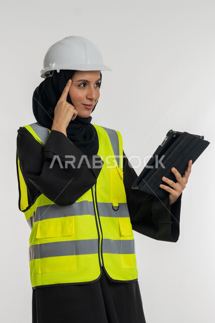 Studying project basics, working in the engineering sector, engineering project management, portrait of an Arab Gulf Emirati female engineer wearing a protective jacket and helmet working on a tablet, a young Saudi woman putting her hand on her head with thinking gestures, white background