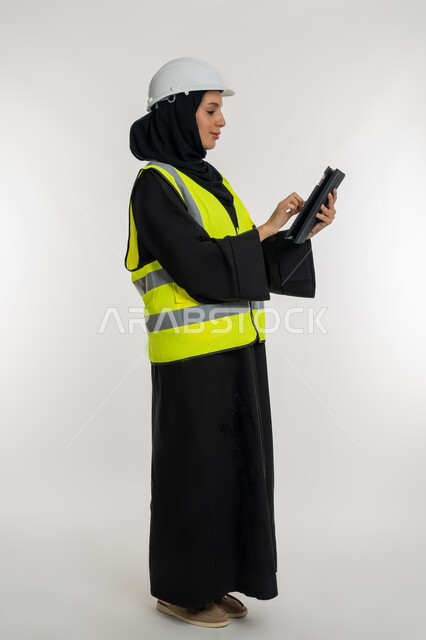 Working in the engineering sector, engineering project management, remote task completion, portrait of the side of an Emirati Arab-Gulf engineer wearing a jacket and protection helmet working on the tablet, a young Saudi woman studying the basics of the project, a picture of the whole body, white background