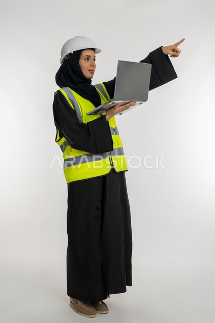 Determine a specific location, work in the engineering sector, accomplishing work tasks remotely, portrait of an Arab Gulf Emirati female engineer wearing a protective jacket and helmet, holding a laptop in her hand, pointing with her index finger to a place, a young Saudi woman studying the basics of the project, a full body image, white background