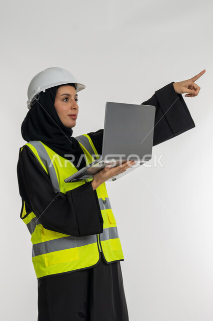 Working in the engineering sector, determining a specific location, completing work tasks remotely, portrait of an Arab Gulf Emirati female engineer wearing a protective jacket and helmet, holding a laptop in her hand, pointing with her index finger to a place, a young Saudi woman studying the basics of the project, engineering project management, white background