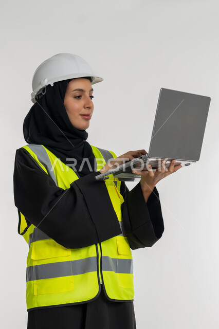 انجاز مهام العمل عبر الحاسوب المحمول, بورتريه لمهندسة عربية خليجية اماراتية ترتدي سترة وخوذة الحماية تمسك الابتوب, ادارة المشاريع الهندسية, العمل في القطاع الهندسي, شابة سعودية تقوم بدراسة أساسيات المشروع, خلفية بيضاء