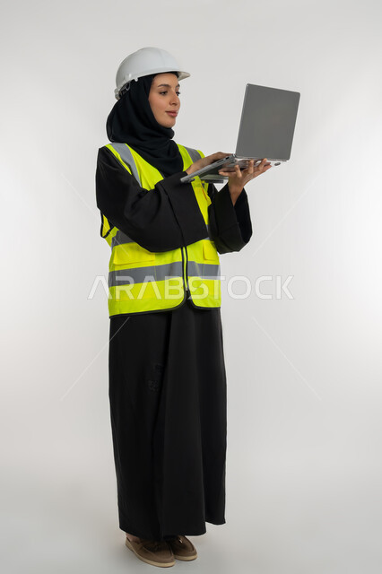 Working in the engineering sector, managing engineering projects, completing work tasks via laptop, portrait of an Arab Gulf Emirati female engineer wearing a protective jacket and helmet holding a laptop, a young Saudi woman studying the basics of the project, a full body image, white background