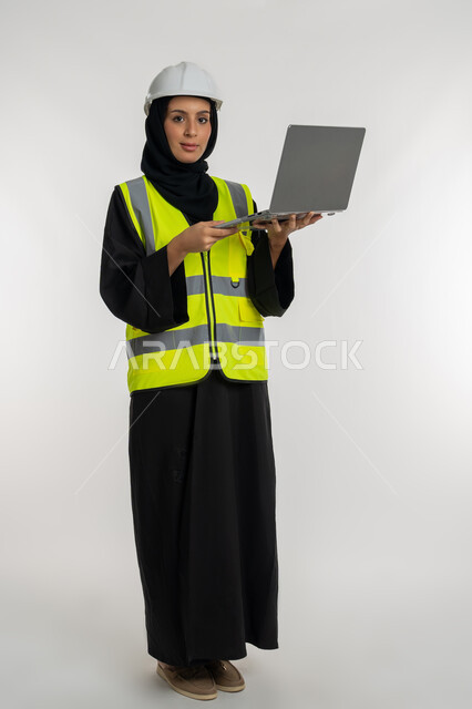 Engineering project management, completing work tasks via laptop, portrait of an Arab Gulf Emirati female engineer wearing a protective jacket and helmet holding a laptop, working in the engineering sector, a young Saudi woman studying the basics of the project, full body image, white background