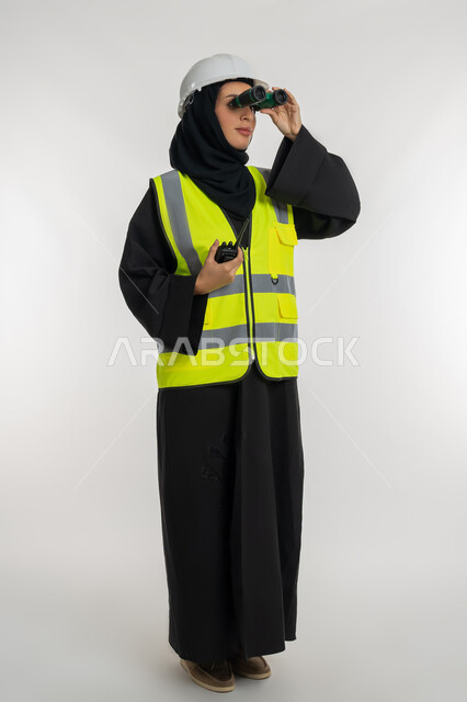 الإشراف على المشاريع، البحث والتخطيط للمستقبل، توسيع مجال الرؤية، العمل في القطاع الهندسي، بورتريه مقرب لمهندسة عربية خليجية اماراتية ترتدي سترة وخوذة الحماية تنظر عبر منظار ثنائي العينين، شابة سعودية تحمل بيدها جهازالاستقبال اللاسلكي، صورة لكامل الجسم، خلفية بيضاء