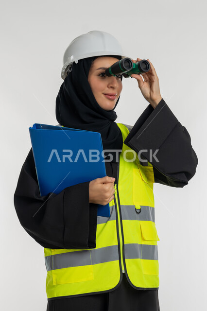 التخطيط والبحث للمستقبل، توسيع مجال الرؤية، الإشراف على المشاريع، العمل في القطاع الهندسي، بورتريه مقرب لمهندسة عربية خليجية اماراتية ترتدي سترة وخوذة الحماية تنظر عبر منظار ثنائي العينين، شابة سعودية تحمل بيدها ملف خاص بالعمل، خلفية بيضاء