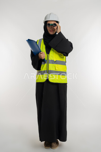 البحث والتخطيط للمستقبل، توسيع مجال الرؤية، الإشراف على المشاريع، العمل في القطاع الهندسي، بورتريه لمهندسة عربية خليجية اماراتية ترتدي سترة وخوذة الحماية تنظر عبر منظار ثنائي العينين، شابة سعودية تحمل بيدها ملف خاص بالعمل، صورة لكامل الجسم، خلفية بيضاء
