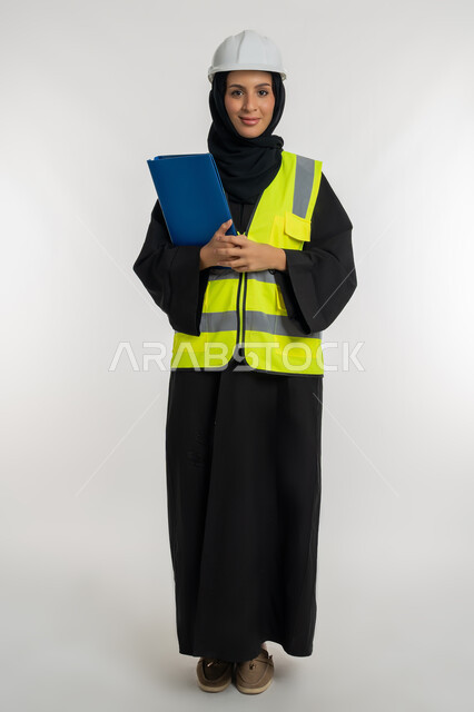 ترتيب وتنظيم البيانات، مهن ووظائف نسائية، الإشراف على المشاريع، بورتريه لمهندسة عربية خليجية اماراتية ترتدي سترة وخوذة الحماية تعمل في القطاع الهندسي، شابة سعودية تحمل بيدها ملف خاص بالعمل، صورة لكامل الجسم، خلفية بيضاء