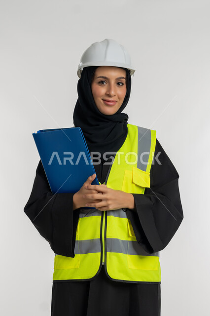 الإشراف على المشاريع، ترتيب وتنظيم البيانات، مهن ووظائف نسائية، بورتريه لمهندسة عربية خليجية اماراتية ترتدي سترة وخوذة الحماية تعمل في القطاع الهندسي، شابة سعودية تحمل بيدها ملف خاص بالعمل، خلفية بيضاء