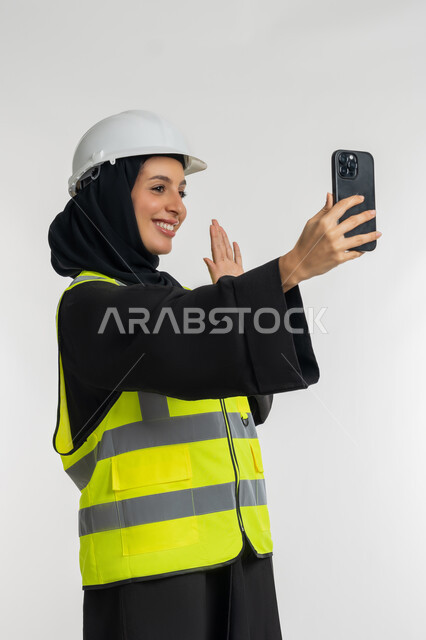 دمج التكنولوجيا الحديثة بالمجال الهندسي، رفع اليد بايماءات الترحيب والقاء التحية، وظائف ومهن نسائية, العمل في القطاع الهندسي, بورتريه من الجانب لمهندسة عربية خليجية امارتية ترتدي خوذة وسترة الحماية تقوم باجراء مكالمة فيديو، شابة سعودية تحمل بيدها الهاتف المحمول، خلفية بيضاء