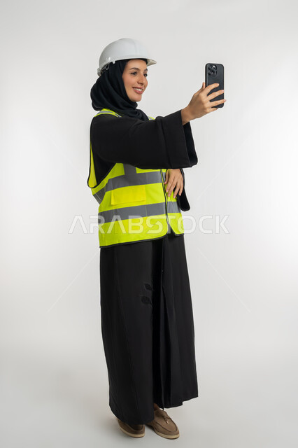 Taking souvenir selfies, integrating modern technology into the engineering field, working in the engineering sector, portrait of an Arab Gulf Emirati female engineer wearing a helmet and a protective vest making a video call, a young Saudi woman holding a mobile phone in her hand, full body photo, white background