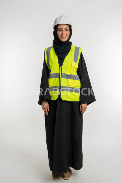 مفهوم إدارة المشاريع الهندسية، وظائف ومهن نسائية, بورتريه لمهندسة معمارية عربية خليجية امارتية ترتدي خوذة وسترة الحماية تقف بتعابير البهجة والسعادة، شابة سعودية تنظر الكاميرا بايماءات السرور تعمل في القطاع الهندسي، صورة لكامل الجسم، خلفية بيضاء
