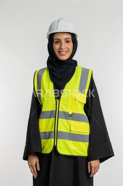 وظائف ومهن نسائية, مفهوم ادارة المشاريع الهندسية, بورتريه لمهندسة معمارية عربية خليجية امارتية ترتدي خوذة وسترة الحماية تقف بتعابير البهجة والسعادة، شابة سعودية تنظر الكاميرا بايماءات السرور تعمل في القطاع الهندسي، خلفية بيضاء