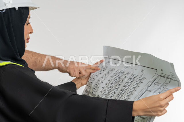 Women's professions and jobs, man pointing to plans and construction projects, working in the engineering sector, close-up portrait from the side of an Arab Gulf Emirati woman wearing a hijab, helmet and protective vest holding a project plan in her hand, a young Saudi woman supervising the work, white background