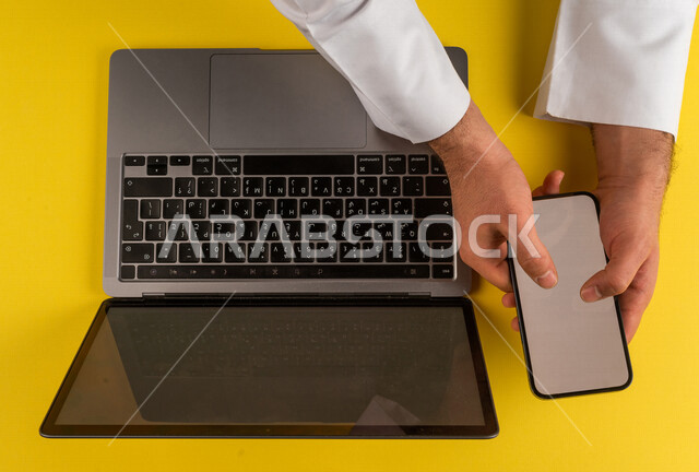 صورة مقربة ليد رجل عربي خليجي سعودي يستخدم الهاتف المحمول وبجانبه الحاسوب المحمول،التسويق للتطبيقات الالكترونية ، نقل البيانات والملفات بين الجهازين، التسوق الالكتروني ، التعليم عن بعد ، استخدام الاجهزة التقنية الحديثة في الاعمال الادارية