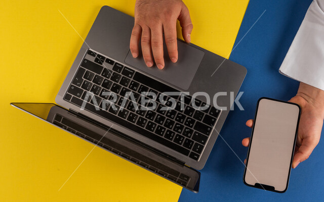 صورة مقربة ليد رجل عربي خليجي سعودي يحمل بيده الهاتف المحمول ويقوم باستخدام الحاسوب المحمول،التسويق والترويج للمواقع الالكترونية نقل البيانات والملفات بين الجهازين، التسوق الالكتروني، التعليم عن بعد ، استخدام الاجهزة التقنية الحديثة في الاعمال الادارية