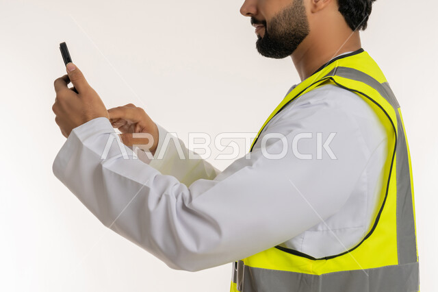 المهن والوظائف الشبابية، العمل في القطاع الهندسي, بورتريه مقرب من الجانب لمهندس عربي خليجي امارتي يرتدي الكندورة وسترة الحماية يقوم بمتابعة سير الأعمال عن بعد عبر الهاتف المحمول، دمج التكنولوجيا المتطورة بالهندسة، الأجهزة التقنية الحديثة، خلفية بيضاء
