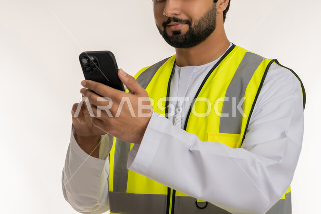 الأجهزة التقنية الحديثة، المهن والوظائف الشبابية في الإمارات، العمل في القطاع الهندسي, بورتريه مقرب لمهندس عربي خليجي امارتي يرتدي الكندورة وسترة الحماية يقوم بمتابعة سير الأعمال عن بعد عبر الهاتف المحمول، دمج التكنولوجيا المتطورة بالهندسة، خلفية بيضاء