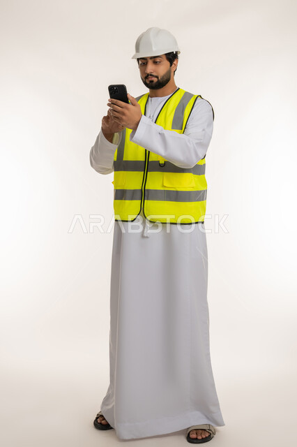 دمج التكنولوجيا الحديثة بالهندسة، المهن والوظائف الشبابية في الإمارات، العمل في القطاع الهندسي, بورتريه لمهندس عربي خليجي امارتي يرتدي الكندورة والخوذة وسترة الحماية يقوم بمتابعة سير الأعمال عن بعد عبر الهاتف المحمول، الجسم بالطول الكامل، خلفية بيضاء