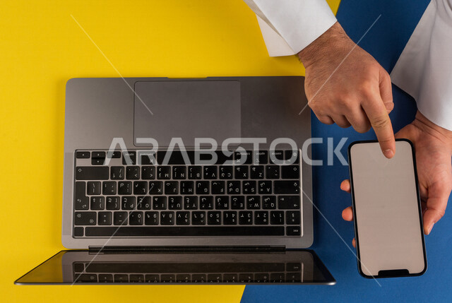 صورة مقربة ليد رجل عربي خليجي سعودي يستخدم الهاتف المحمول وبجانبه الحاسوب المحمول،التسويق للتطبيقات الالكترونية ، نقل البيانات والملفات بين الجهازين، التسوق الالكتروني ، التعليم عن بعد ، استخدام الاجهزة التقنية الحديثة في الاعمال الادارية