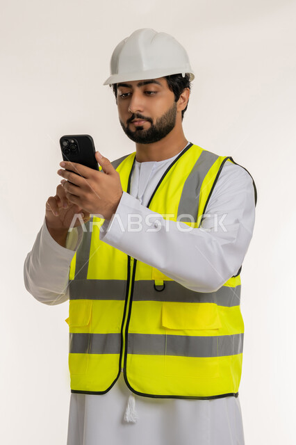 المهن والوظائف الشبابية في الإمارات، العمل في القطاع الهندسي, بورتريه مقرب لمهندس عربي خليجي امارتي يرتدي الكندورة والخوذة وسترة الحماية يقوم بمتابعة سير الأعمال عن بعد عبر الهاتف المحمول، دمج التكنولوجيا المتطورة بالهندسة، الأجهزة التقنية الحديثة، خلفية بيضاء