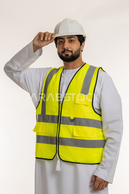 مفهوم إدارة المشاريع الإعمارية، المهن والوظائف الشبابية في الإمارات العربية المتحدة، بورتريه لمهندس عربي خليجي إماراتي يرتدي الكندورة والخوذة وسترة الحماية يقف باستقامة ينظر إلى الكاميرا بإيماءات الجدية والثقة، العمل في القطاع الهندسي، خلفية بيضاء