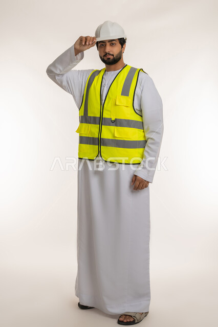 Working in the engineering sector, managing construction projects, youth professions and jobs in the United Arab Emirates, portrait of an Arab Gulf Emirati engineer wearing a kandura, helmet and protective vest, standing straight, looking at the camera with gestures of seriousness and confidence, full body, white background