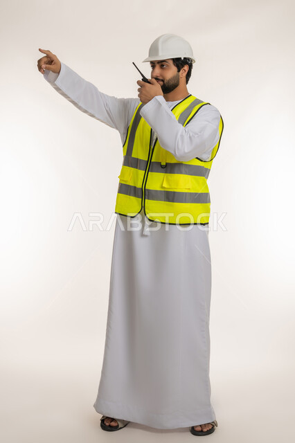 إدارة المشاريع الهندسية الإعمارية، بورتريه لمهندس عربي خليجي إماراتي يرتدي الكندورة والخوذة وسترة الحماية يحمل جهاز اتصال لاسلكي يشير بإصبع السبابة إلى شيء ما في الأعلى، المهن والوظائف الشبابية، إعطاء أوامر خاصة بالعمل، صورة الجسم بالطول الكامل، خلفية بيضاء
