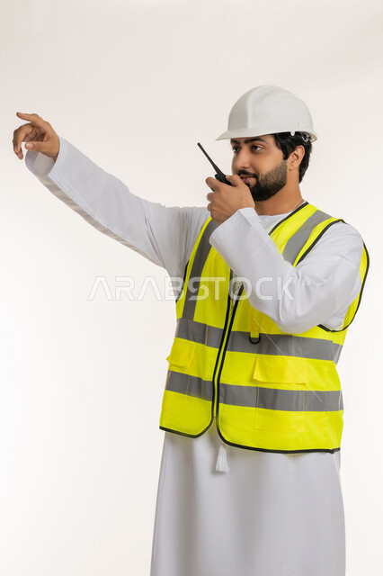 إعطاء أوامر خاصة بالعمل، إدارة المشاريع الهندسية الإعمارية، بورتريه لمهندس عربي خليجي إماراتي يرتدي الكندورة والخوذة وسترة الحماية يحمل جهاز اتصال لاسلكي يشير بإصبع السبابة إلى شيء ما في الأعلى، المهن والوظائف الشبابية، خلفية بيضاء