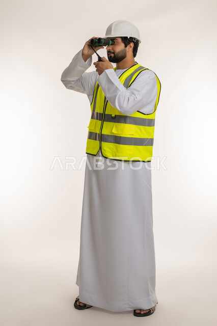 Youth jobs and professions, following up on engineering projects, seeing distant objects, portrait of an Arab Gulf Emirati engineer wearing a kandura, helmet and protective vest, holding a wireless communication device, looking through binoculars, giving work orders, full-length body image, white background