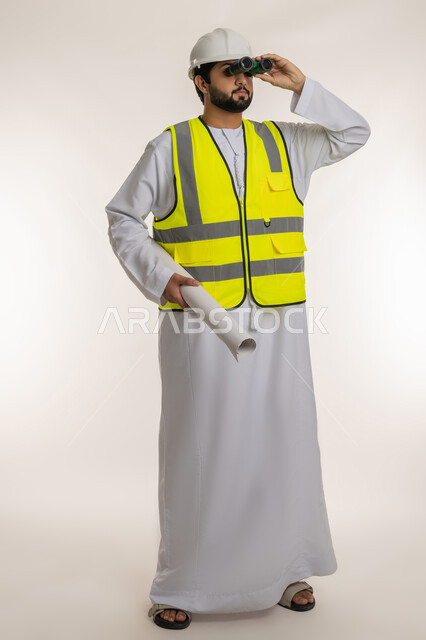 Seeing distant objects, portrait of an Arab Gulf Emirati engineer wearing a kandura, helmet and protective vest looking through binoculars at something holding a blueprint in his hand, following the progress of construction engineering projects, youth professions and jobs, full-length body image, white background