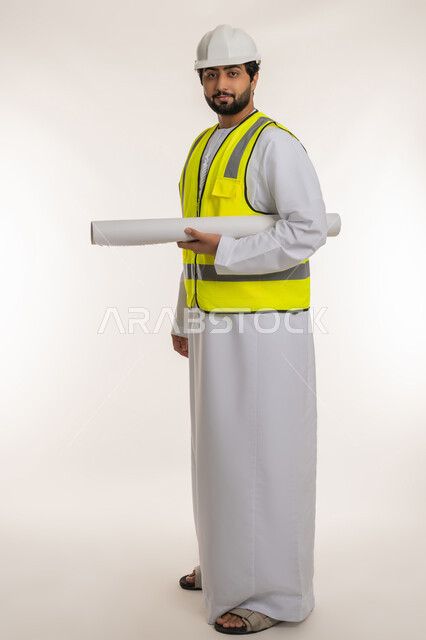 Concept of architectural construction and engineering, portrait of an Arab Gulf Emirati engineer wearing a kandura, helmet and protective vest holding a blueprint looking at the camera with gestures of confidence, following up on the progress of construction projects, youth professions and jobs, full body image, white background