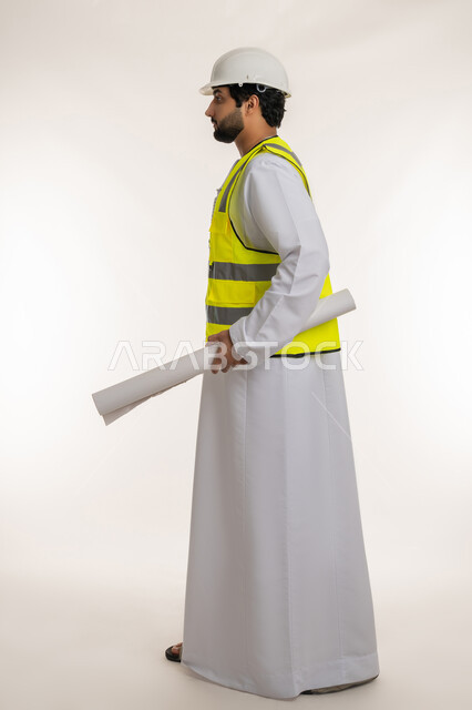 مفهوم الإنشاءات المعمارية والهندسة، بورتريه من الجانب لمهندس عربي خليجي إماراتي يرتدي الكندورة والخوذة وسترة الحماية يحمل بيده مخطط ينظر إلى شيء ما، المهن والوظائف الشبابية في الإمارات، متابعة سير المشاريع الإعمارية، الجسم بالطول الكامل، خلفية بيضاء