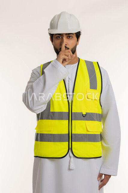 طلب السكوت والهدوء، الأمر بالتوقف عن الكلام، مفهوم الإنشاءات المعمارية، بورتريه لمهندس عربي خليجي إماراتي يرتدي الكندورة والخوذة وسترة الحماية يضع إصبع السبابة فمه، العمل في القطاع الهندسي، المهن والوظائف الشبابية في الإمارات العربية المتحدة، خلفية بيضاء