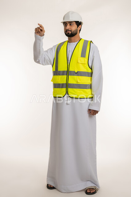 العمل في القطاع الهندسي، المهن والوظائف الشبابية في الإمارات، مفهوم الإنشاءات المعمارية، بورتريه لمهندس عربي خليجي إماراتي يرتدي الكندورة والخوذة وسترة الحماية يشير بإصبع السبابة إلى شيء ما، الجسم بالطول الكامل، خلفية بيضاء