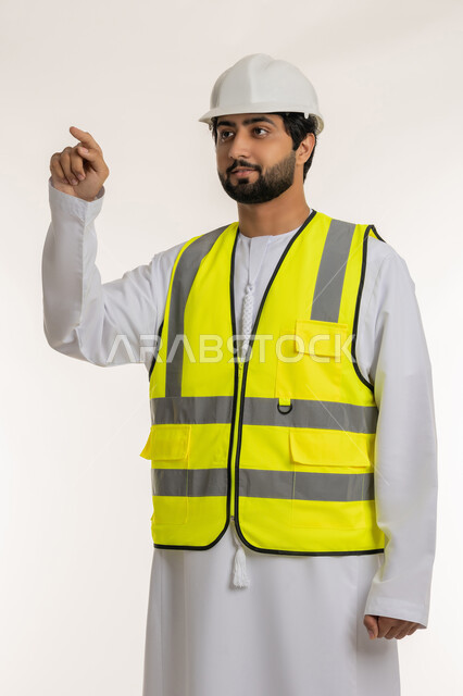 مفهوم الإنشاءات المعمارية، بورتريه لمهندس عربي خليجي إماراتي يرتدي الكندورة والخوذة وسترة الحماية يشير بإصبع السبابة إلى شيء ما، العمل في القطاع الهندسي، المهن والوظائف الشبابية في الإمارات العربية المتحدة، خلفية بيضاء