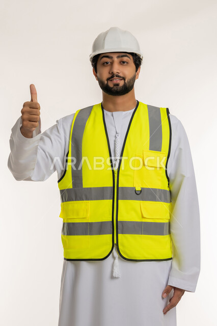 مفهوم الإنشاءات المعمارية، بورتريه لمهندس عربي خليجي إماراتي يرتدي الكندورة والخوذة وسترة الحماية يرفع إصبع الإبهام للأعلى بإيماءات الإعجاب، علامة الجودة والامتياز، المهن والوظائف الشبابية في الإمارات العربية المتحدة، خلفية بيضاء