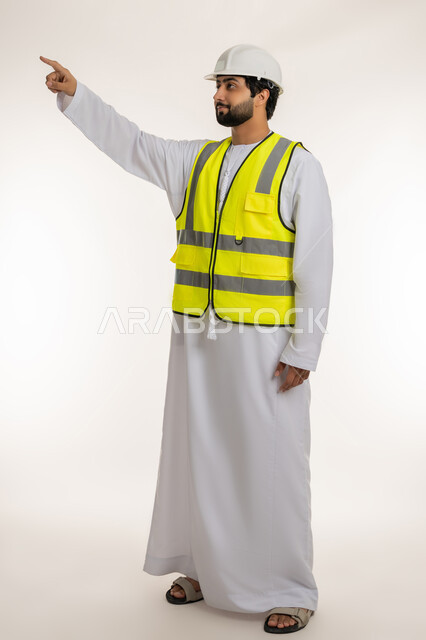 Youth jobs and professions in the Emirates, concept of architectural construction and engineering, portrait of an Arab Gulf Emirati engineer wearing a kandura, helmet and protective vest pointing with index finger to something above, full body image, white background