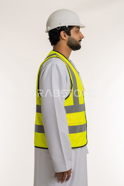 Concept of architectural construction and engineering, side portrait of an Arab Gulf Emirati engineer wearing a kandura, helmet and protective vest standing straight looking at something, youth professions and jobs in the Emirates, white background