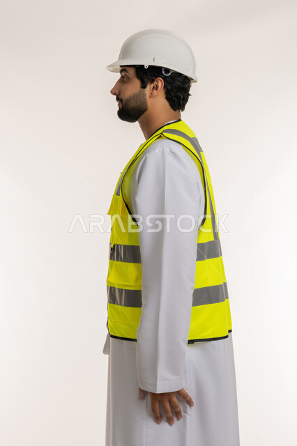 Working in the engineering sector, concept of architectural construction and engineering, side portrait of an Arab Gulf Emirati engineer wearing a kandura, helmet and protective vest standing straight looking at something, youth professions and jobs in the Emirates, white background
