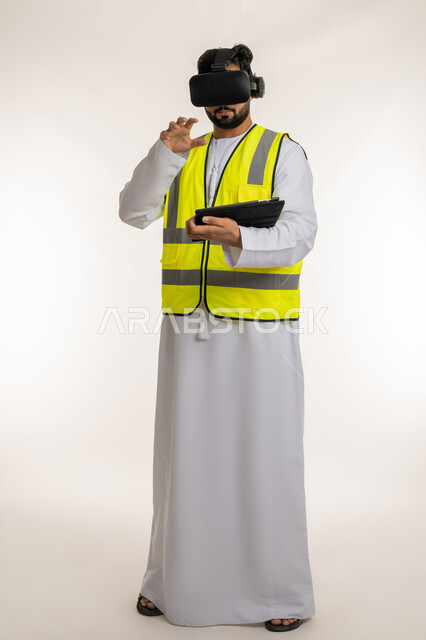 التفاعل مع الهندسة الافتراضية، دمج التكنولوجيا في المجال الهندسي في الإمارات، بورتريه لمهندس عربي خليجي إماراتي يرتدي الكندورة والسترة الخاصة بالعمل ونظارة الواقع الافتراضي يرفع يده للمس عناصر هولوجرامية يمسك الجهاز اللوحي، الجسم بالطول الكامل، خلفية بيضاء