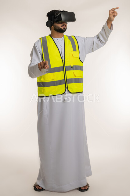 تجسيد ومشاهدة المشاريع الهندسية بالاعتماد على التكنولوجيا، الاندماج مع الهندسة الافتراضية، بورتريه لمهندس عربي خليجي إماراتي يرتدي الكندورة والسترة الخاصة بالعمل ونظارة الواقع الافتراضي يرفع يديه للمس عناصر هولوجرامية، الجسم بالطول الكامل، خلفية بيضاء