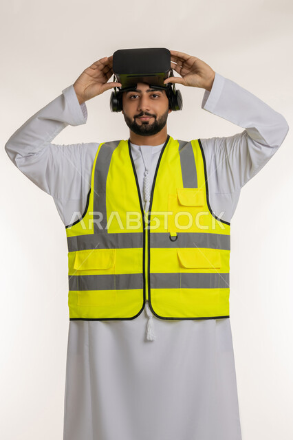 Integrating technology with engineering fields, development of engineering field, interaction with virtual engineering, portrait of an Arab Gulf Emirati engineer wearing a kandura and a work jacket, putting virtual reality glasses on his head, looking at the camera with gestures of confidence, future vision 2030, white background